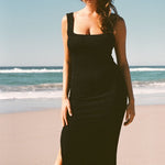 A woman on the beach wearing black textured bodycon midi dress with inbuilt bra support, square neckline, side split. 