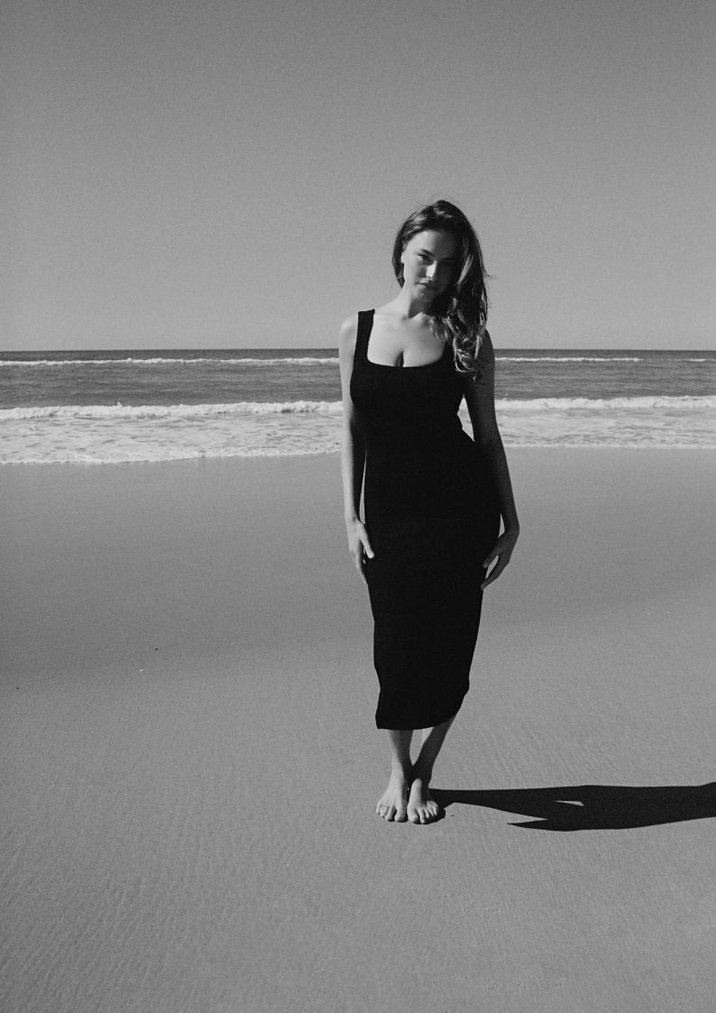 A woman on the beach wearing black textured bodycon midi dress with inbuilt bra support, square neckline, side split. 