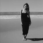A woman on the beach wearing black textured bodycon midi dress with inbuilt bra support, square neckline, side split. 