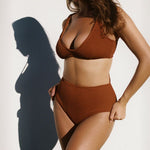 Women wearing brown rust caramel D+ bikini top and high waisted bikini bottom in textured fabric against white wall. 