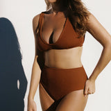 Women wearing brown rust caramel D+ bikini top and high waisted bikini bottom in textured fabric against a white wall. 